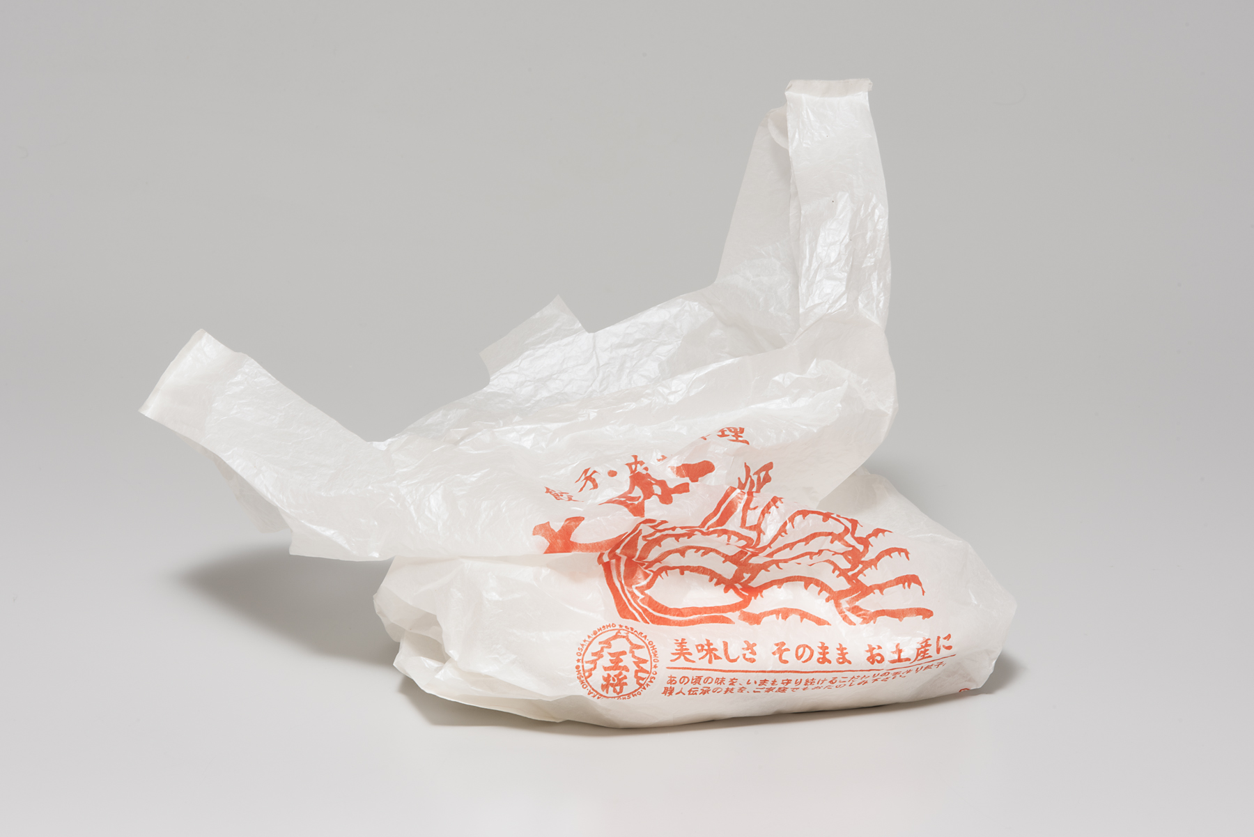 This porcelain piece is constructed to look identical to a white plastic bag. The bag is slightly crumpled and holds a porcelain version of a Styrofoam takeout container. The front of the white bag has red text in Japanese and the logo of Osaka Ohsho, the restaurant this piece is modeled after.