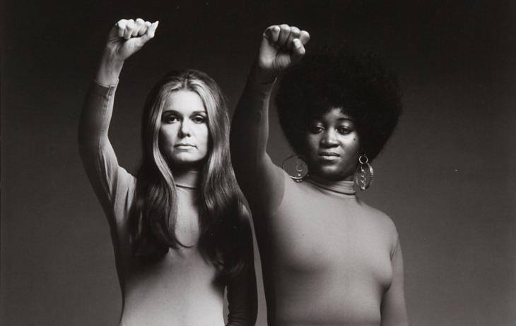 Gloria Steinem and Dorothy Pitman Hughes stand side by side, each raising their right fist into the air