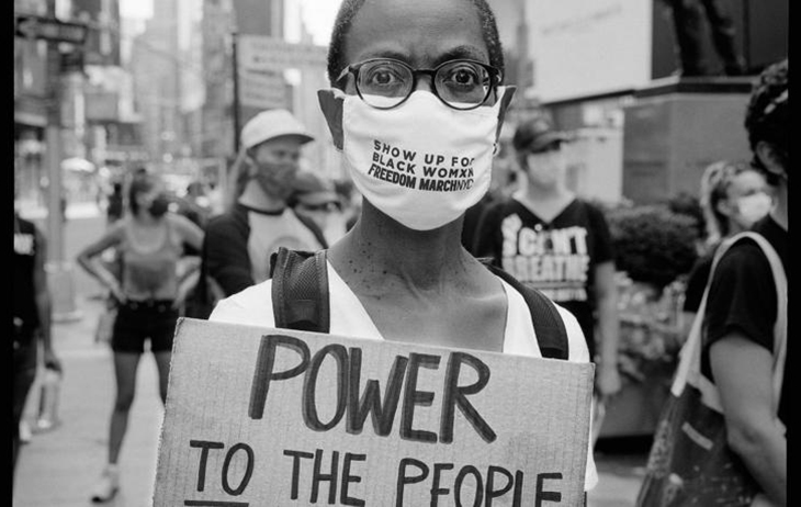 A Black woman with close-cropped hair wearing glasses and a face mask that reads “SHOW UP FOR BLACK WOMXN FREEDOM MARCHNYC” holds a hand-lettered cardboard sign reading “POWER TO THE PEOPLE”