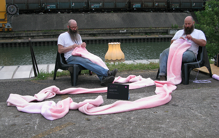 Two white men with long beards sit outside crocheting with pink yarn