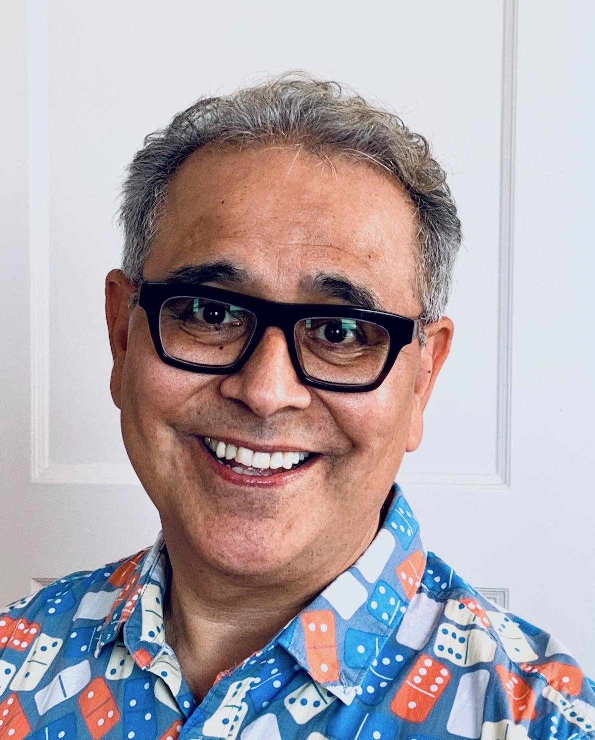 Photographic color portrait of a man. He is wearing thick framed glasses and a brightly colored shirt and smiling directly at the camera.