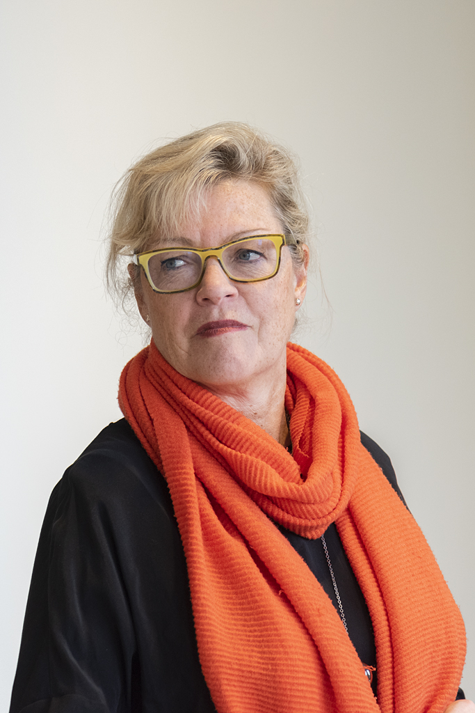 Photographic color portrait of a light-skinned woman with blonde hair, yellow glasses, and a bright orange scarf