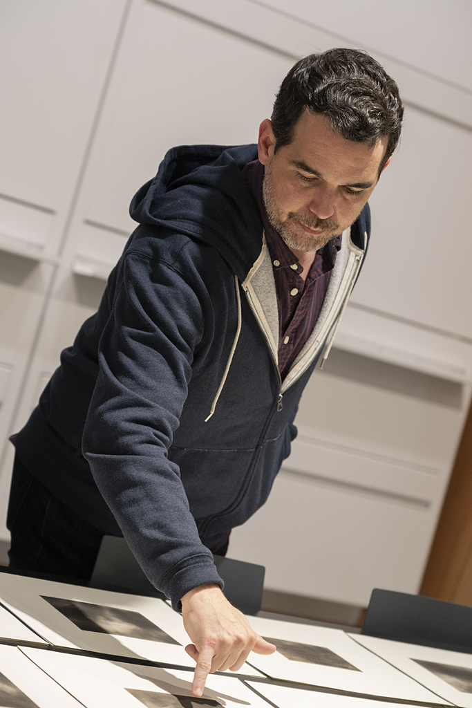Photograph of Dario Robleto, a dark haired man in a hoodie, looking at prints on a table