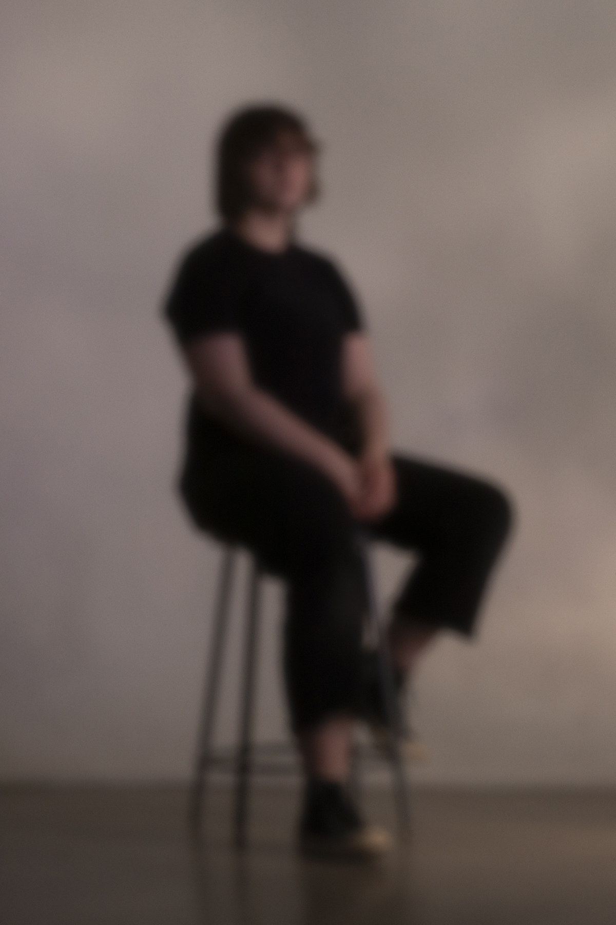 A person dressed in all black sits on a stool with their hands in their lap. The image is out of focus.