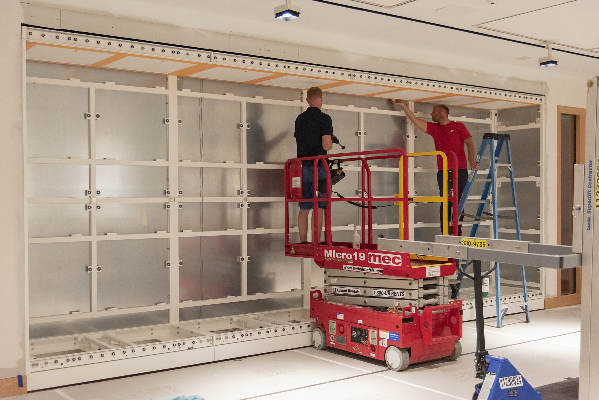 Two workers stand on a lift while installing casing in a bright white room