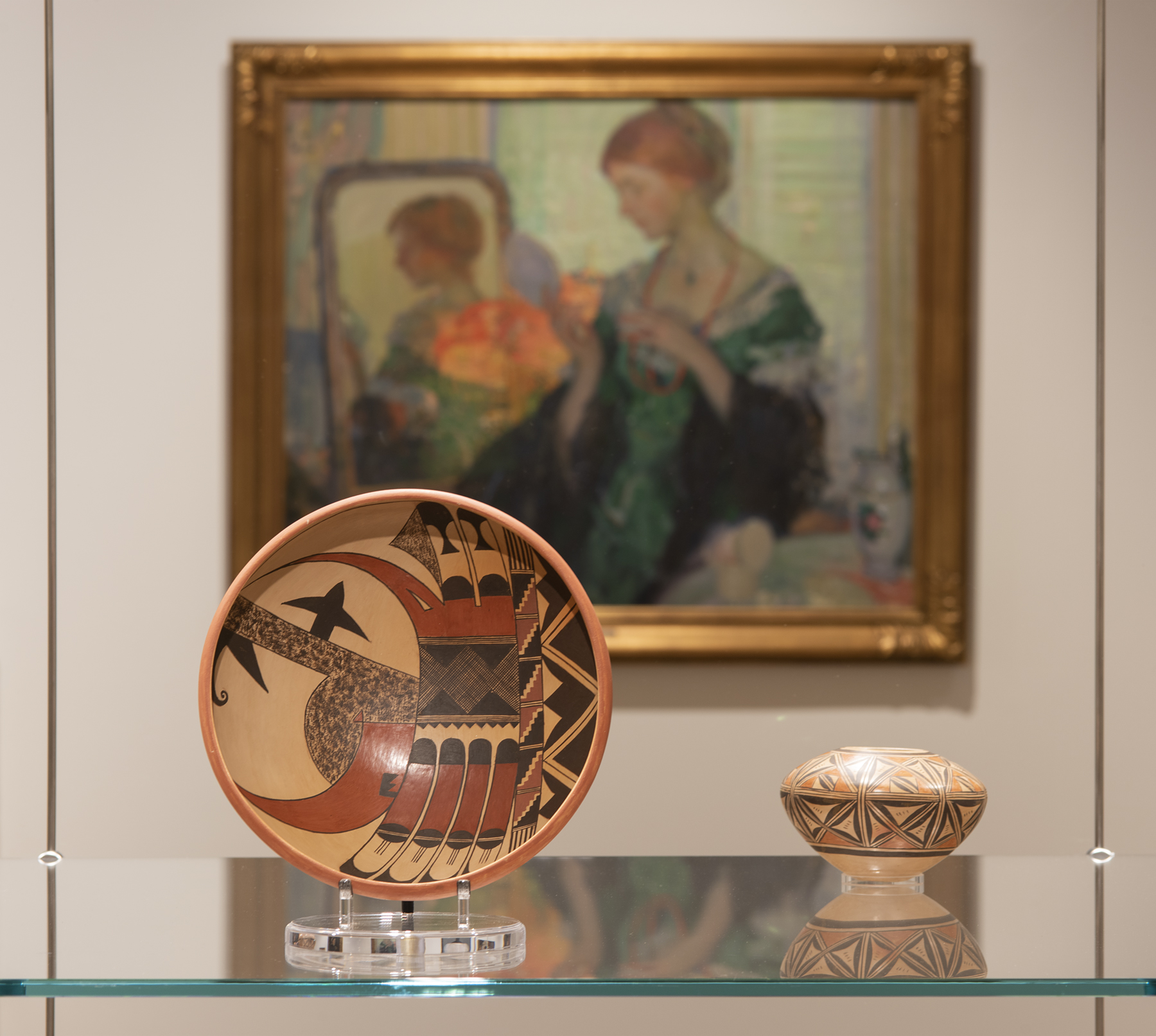 Two small ceramic objects sit on a glass shelf with a painting visible in the background