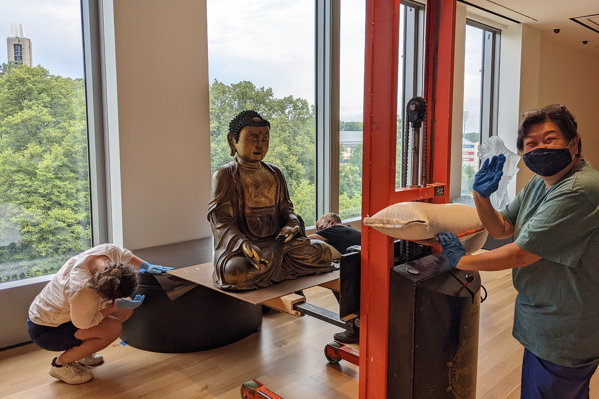Three people working in front of a window move a Buddha sculpture using machinery