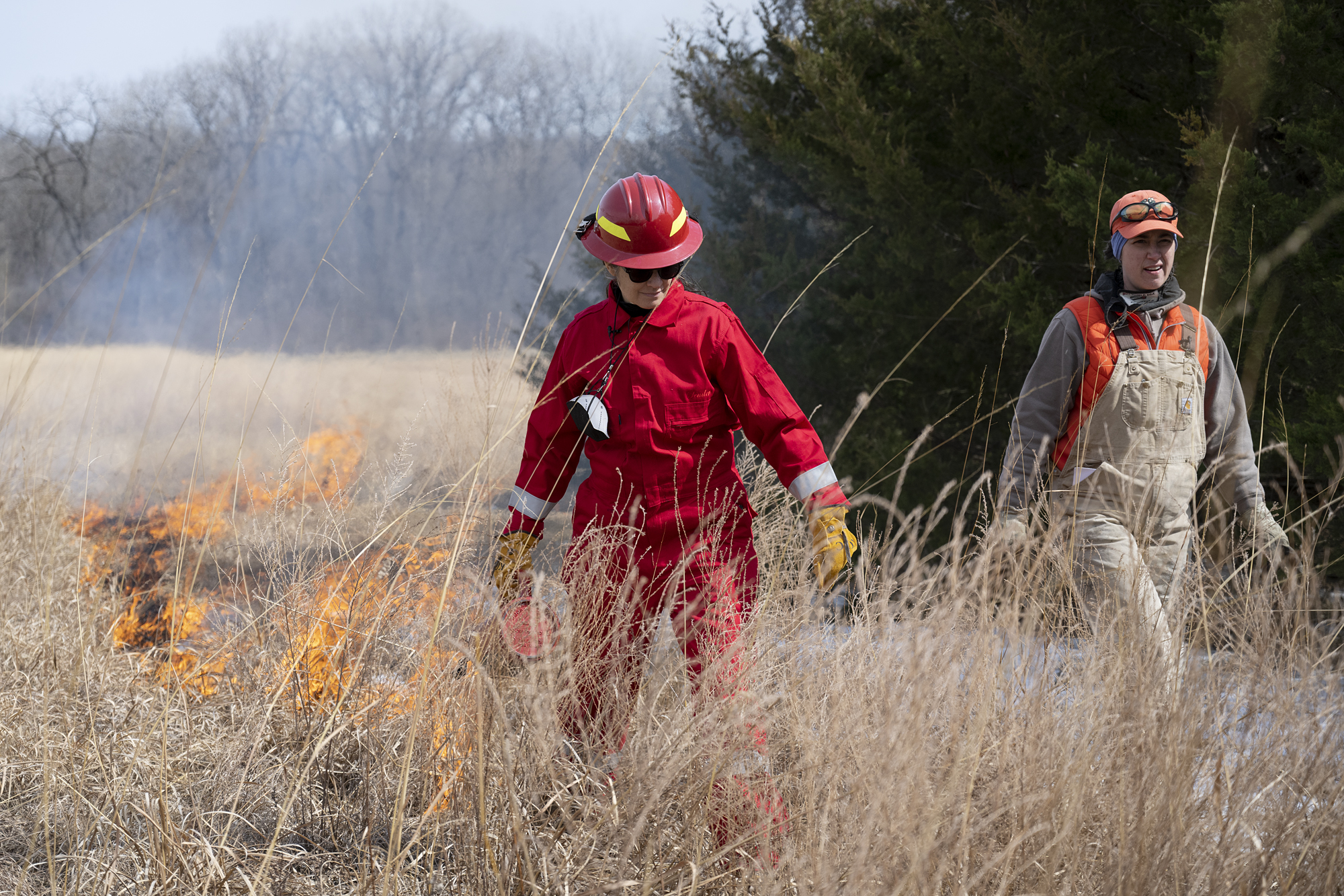 A woman in a red fire suit and red hard hat ignites a field of brown grass while behind her the field burns