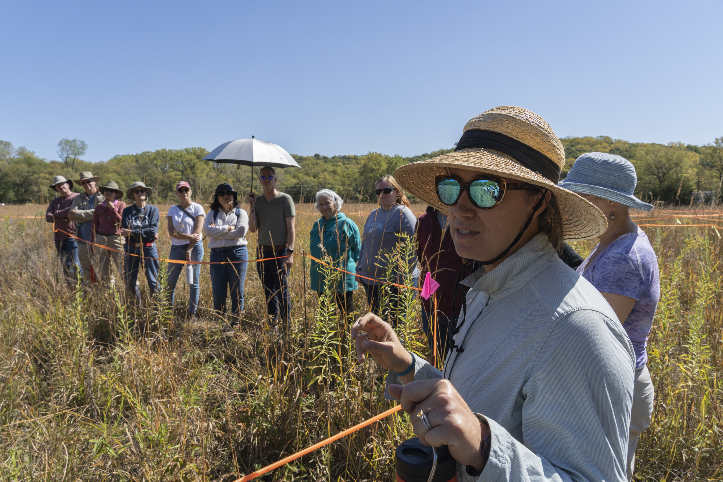 A white woman in a hat and sunglasses talks to a group of people in a field