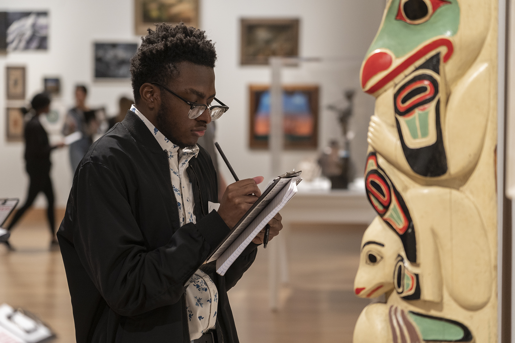 A student with glasses holds a clipboard and takes notes while standing in front of a totem pole