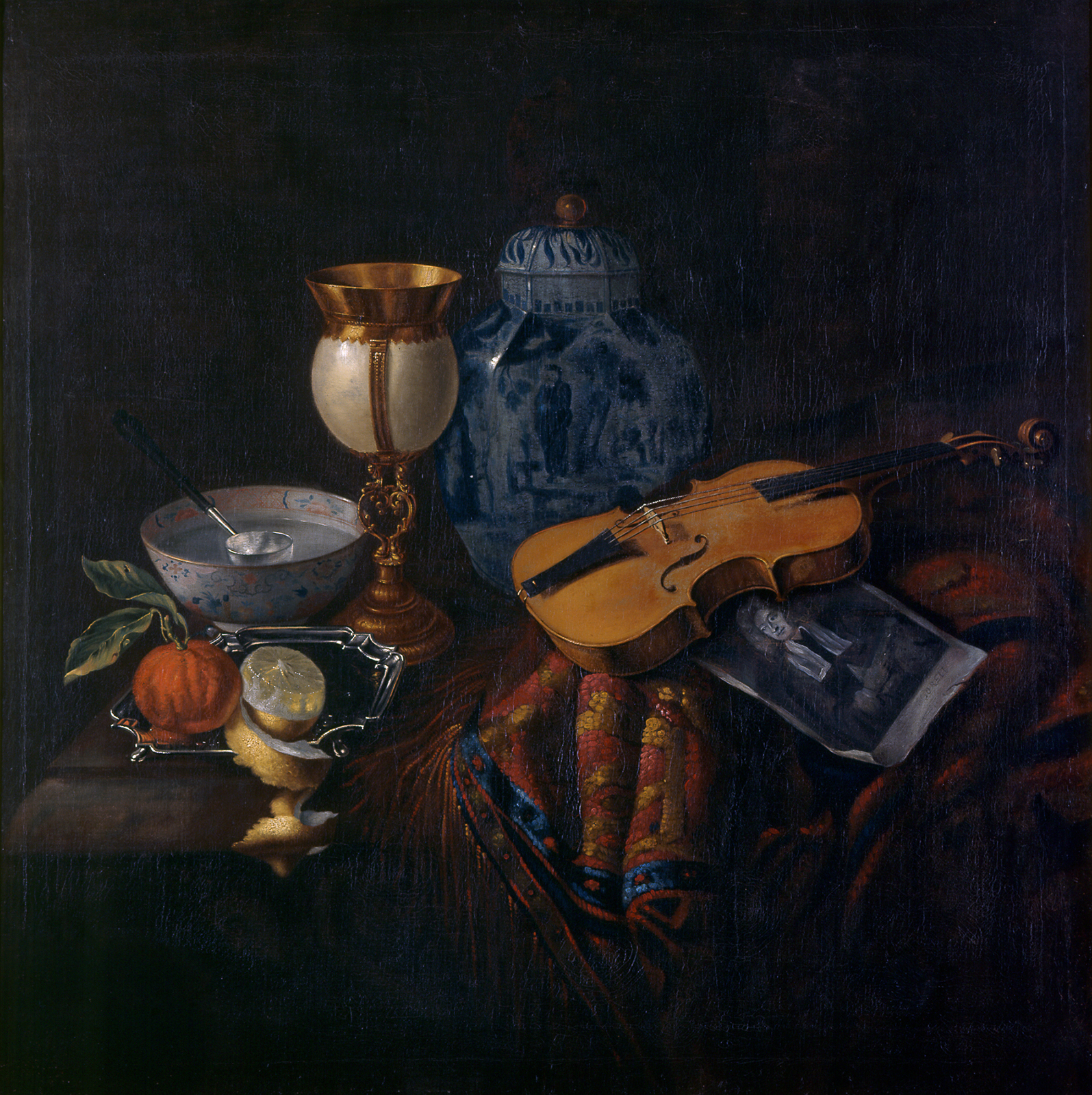 Items arranged on a table include a peeled lemon and an orange, a bowl of water, a gold-and-white goblet, a blue-and-white urn, a violin, and a portrait of a white man wearing a wig