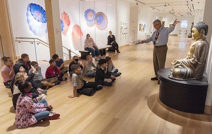 A group of children sit on the floor in a gallery listening to an older man talk as he gestures to a sculpture