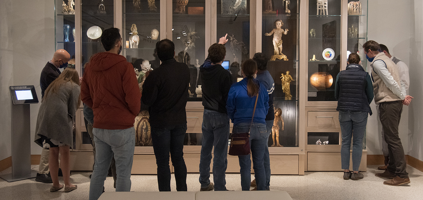 Ten visitors stand in front of a glass cabinet with many objects on view.