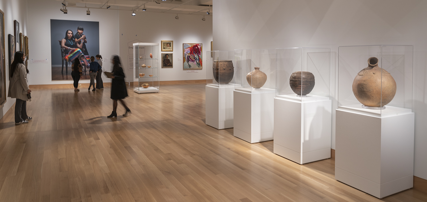 A bright gallery with four large pots lining the right-hand wall and visitors view works of art in the background