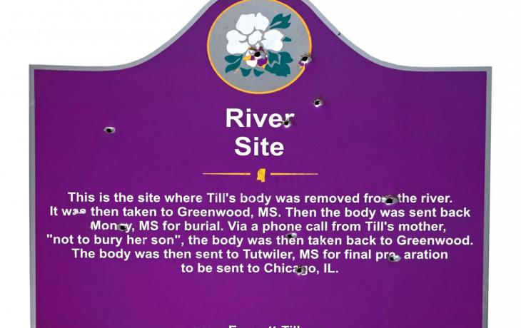 purple sign with bullet holes and a paragraph of white text that begins "River Site: This is the site where Emmett Till's body was removed from the river."