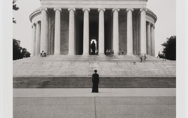 This black-and-white photograph shows a solitary woman identified as the artist, Carrie Mae Weems, standing at the bottom of the stone steps leading to the Thomas Jefferson Memorial in Washington, DC. Her back is to the viewer and her stance mimics that of the statue seen at the top of the steps behind the columns of the monument. 