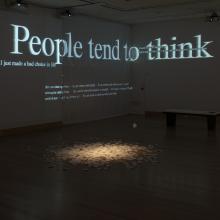 a small pile of white receipt papers grows in the middle of a dark gallery room with words projected in white on the walls
