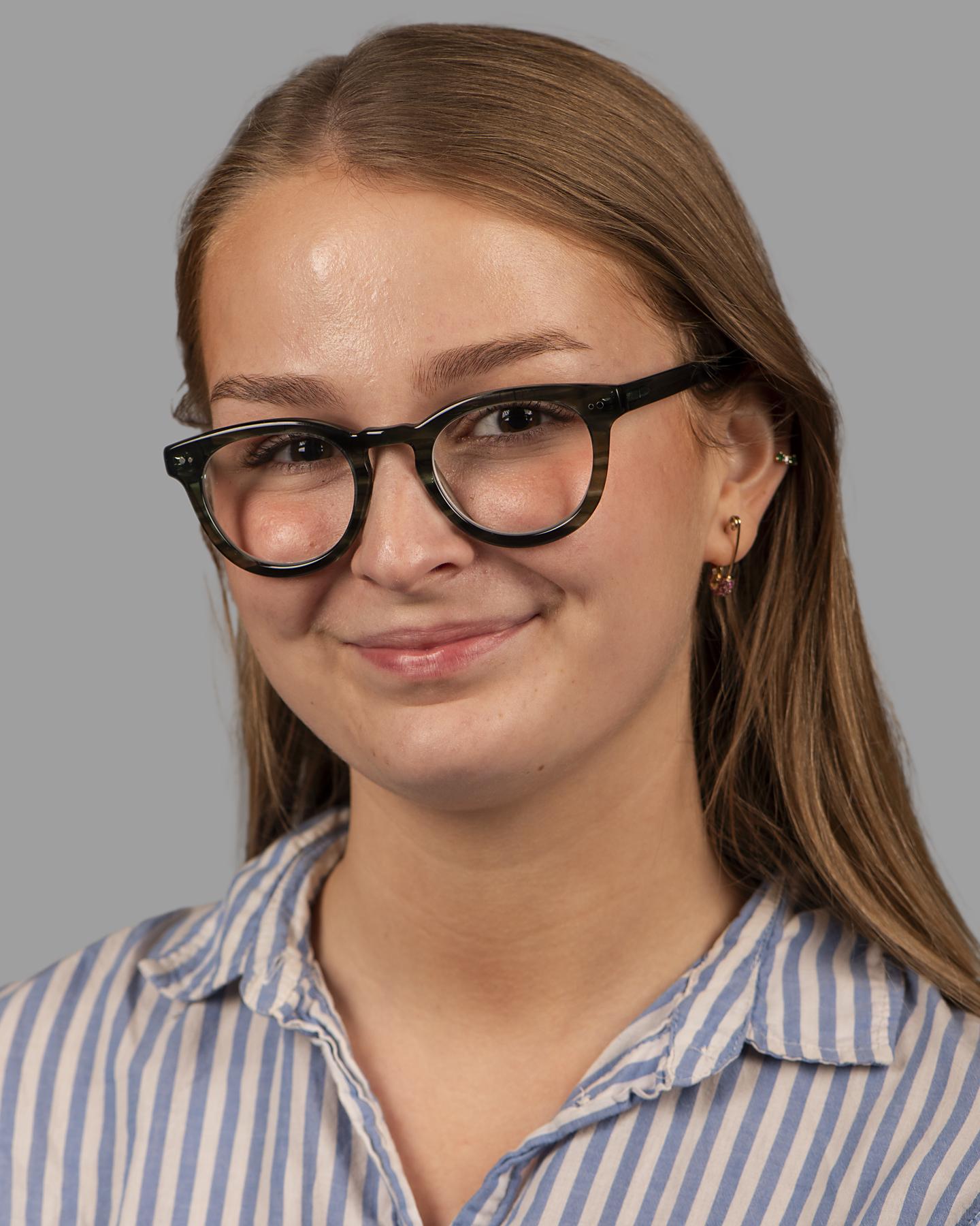 A white woman with long blonde hair wears black circle framed glasses and smiles at the camera