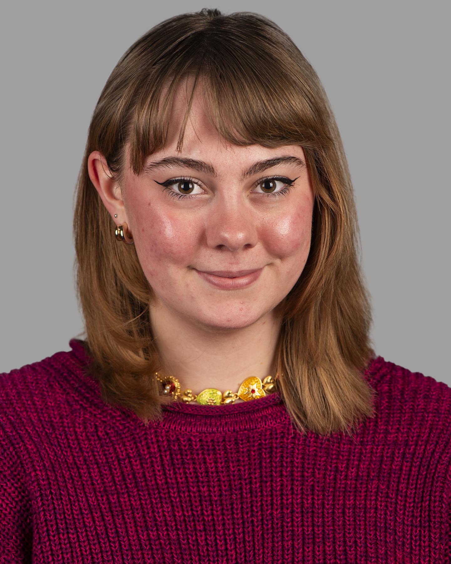 A white woman with shoulder-length brown hair and bangs wears a bright red sweater and chunky gold necklace; she smiles at the camera