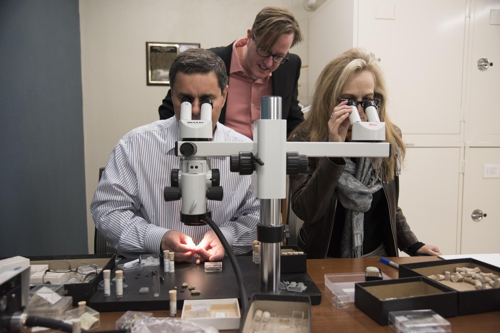 A man and a woman sit in a lab and look into the eyepieces of a large microscope, a man stands over them