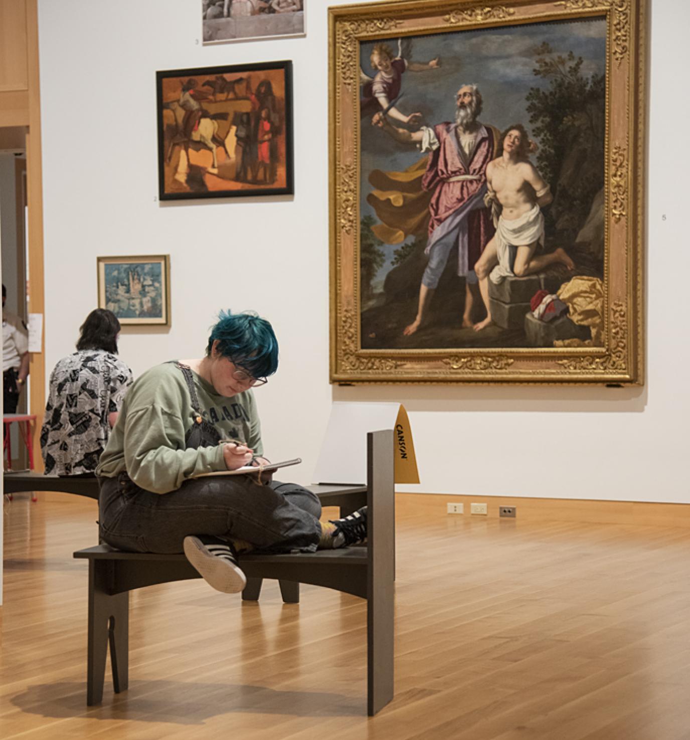 A young adult with short green-blue hair sits in the middle of an art gallery holding a sketchbook
